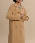 Someone wears the Olivia Coat—an essential oversized camel wool outerwear piece—over a green sweater, standing against a beige background. Their hand holds the coat closed, with their face partially out of frame.