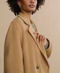 A woman with curly hair, gold hoop earrings, and a ring stands against a neutral background wearing the Olivia Coat—an oversized camel wool coat—layered over a green sweater. Her upper body and face are partially visible.