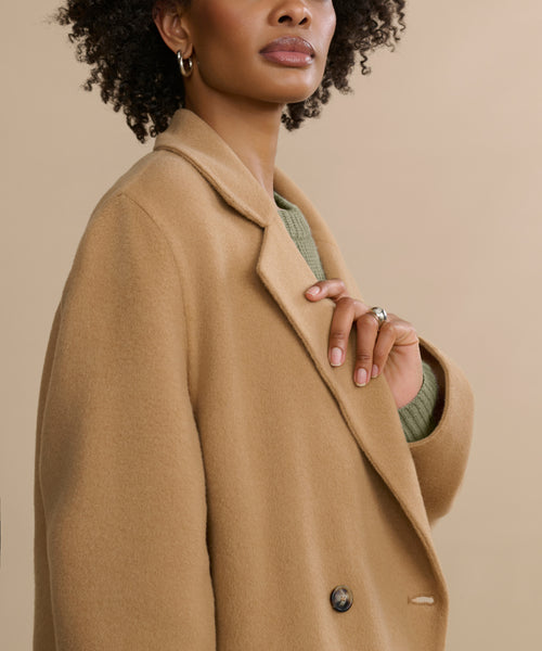 A woman with curly hair, gold hoop earrings, and a ring stands against a neutral background wearing the Olivia Coat—an oversized camel wool coat—layered over a green sweater. Her upper body and face are partially visible.
