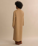 A person with curly hair, seen from behind, wears the Olivia Coat—an oversized camel wool piece—over brown pants against a plain beige background. An outerwear essential for timeless style.