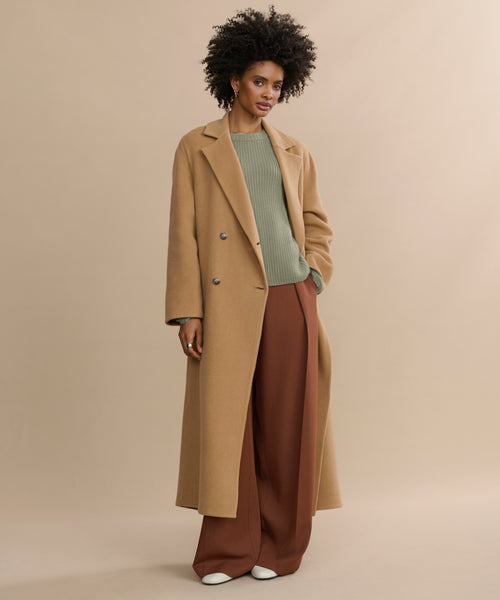 A woman with curly hair poses confidently against a beige background, wearing the oversized Olivia Coat in camel wool over a green sweater, wide brown trousers, and white shoes, with one hand casually placed in her pocket.