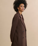 A woman with curly hair wears the Olivia Coat, a long, chocolate double-breasted style, standing against a beige background and gazing confidently at the camera.