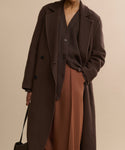 Wearing the oversized Olivia Coat in dark brown over a matching sweater and loose rust-colored pants, the individual holds a dark bag against a beige background, with their face not shown.