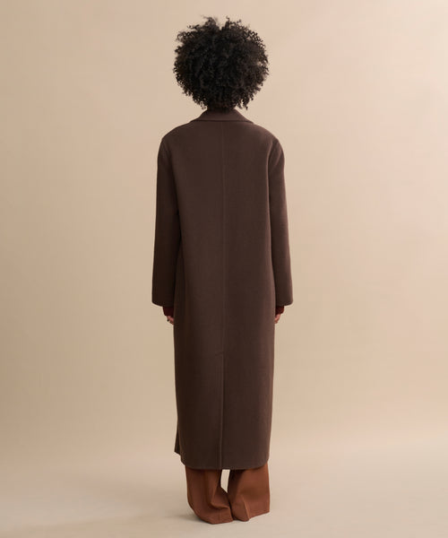 A person stands with their back to the camera, wearing the oversized Olivia Coat in wool chocolate and light brown pants against a plain beige background.