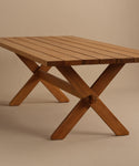 The Outdoor Garden Dining Table features a teak slatted top and crossed X-shaped legs, shown against a plain beige background.