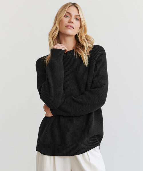 A woman with long blonde hair wears the Oversized Cotton Fisherman sweater in black and white pants, standing against a plain light background. She faces the camera with a relaxed expression, her left hand gently touching her shoulder.
