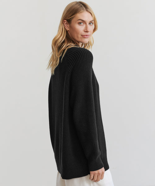 A woman with long blonde hair models the Oversized Cotton Fisherman sweater in black, posing slightly turned to the side against a plain light background.