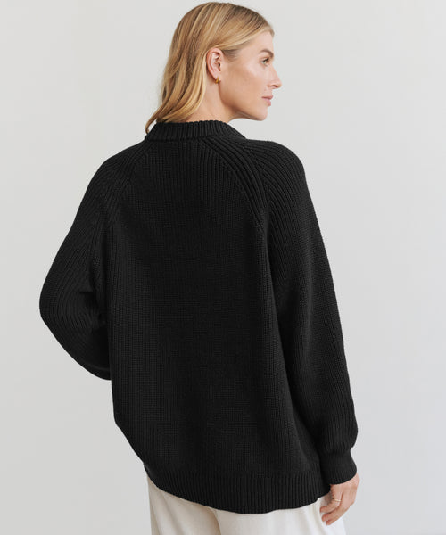 A blonde woman wears the Oversized Cotton Fisherman sweater in black with light pants, standing slightly to the side against a plain light background.