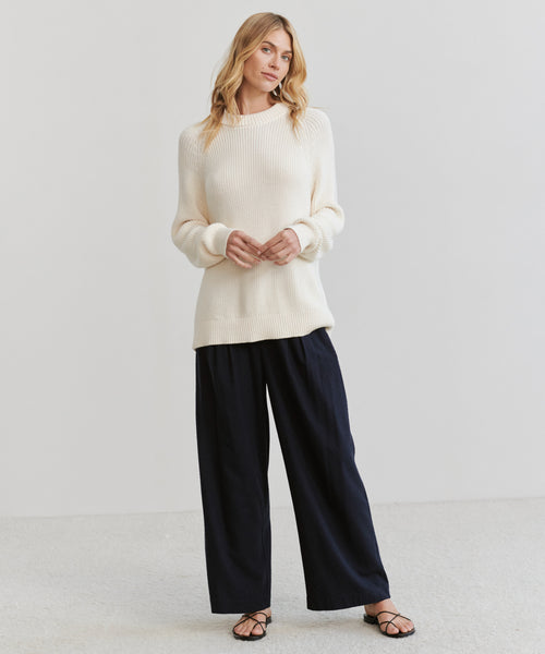 A woman with wavy blonde hair poses against a plain background wearing the Oversized Cotton Fisherman sweater, loose black pants, and black strappy sandals, her hands gently clasped.