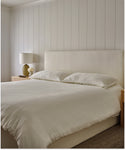 The Pacific Bed with white bedding and an upholstered headboard stands in a minimalist bedroom, accompanied by a wooden nightstand, table lamp, and small vase against a white paneled wall.