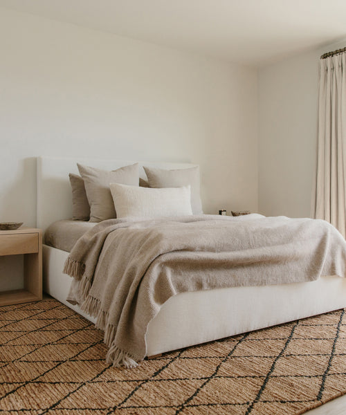 A minimalist bedroom with the Pacific Bed in ivory linen upholstery, beige bedding, and layered pillows. A wooden nightstand sits to the left, beige curtains frame the window, and a patterned woven rug covers the floor.