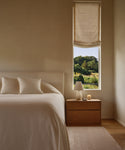 A minimalist bedroom showcases the Pacific Bed in ivory wool boucle with an upholstered headboard, cream pillows, a wooden nightstand topped with a white lamp and candle, and a tall window framed by partial beige curtains revealing lush greenery.