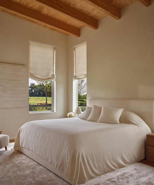 A cozy bedroom features the Pacific Bed with an upholstered headboard, dressed in cream linens and two pillows. A soft rug, light wood beams, matching furniture, and Roman-shaded windows enhance the warmth and offer greenery views.