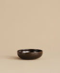 A small, round, dark brown Pacific Breakfast Bowl sits empty and centered on a beige surface with a plain beige background, highlighting its elegant simplicity.