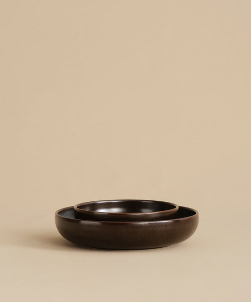 Two dark brown Pacific Soup Bowls, with a smaller one nested inside a larger, rest on a beige surface and matching background. Their minimalist arrangement highlights the elegant design of this dinnerware set.