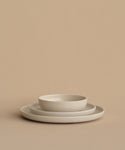 Three minimalist, off-white ceramic plates are stacked on a beige surface. The smallest plate is the Pacific Breakfast Bowl, a shallow stoneware piece, resting atop two larger matching flat plates.