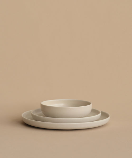 Three minimalist, off-white ceramic plates are stacked on a beige surface. The smallest plate is the Pacific Breakfast Bowl, a shallow stoneware piece, resting atop two larger matching flat plates.
