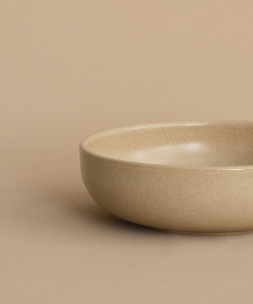 A partial view of the Pacific Breakfast Bowl, a shallow and round beige stoneware piece, is shown on a matching beige surface against a plain beige background, positioned toward the right side of the image.