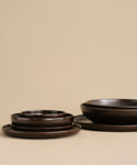 The Pacific Dinner Plate, crafted from dark brown ceramic stoneware, is showcased with matching bowls against a beige background. The minimalist display emphasizes the smooth, matte texture of this elegant dinnerware set.