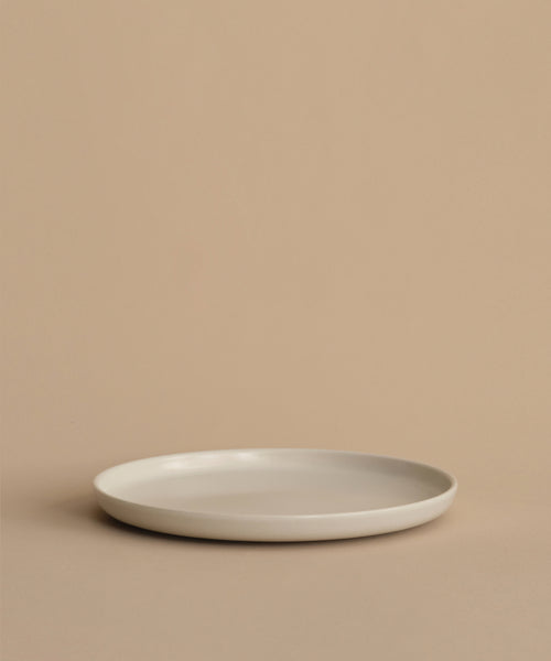 The Pacific Dinner Plate, a simple round white stoneware piece, rests on a beige surface against a matching beige background for a minimalist and neutral look.