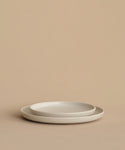 Two simple, round Pacific Dinner Plates in white stoneware are stacked against a beige background. Their minimalist design and soft lighting create a clean, modern look for any setting. Made in Portugal.