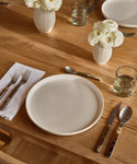 A simple table setting on a wooden table includes a Pacific Dinner Plate, silverware, a glass of water, a white napkin, and two white flower arrangements in vases.