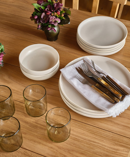 A wooden table set with stacked Pacific Dinner Plates, bowls, four glasses, cutlery wrapped in a white napkin, and two small vases with purple flowers.
