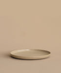The Pacific Dinner Plate, a simple round plate made from beige ceramic, sits on a matching surface with a plain beige backdrop.