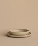 Two round Pacific Dinner Plates are stacked on a matching beige surface and background, with the smaller, deeper plate resting inside the larger, flatter Pacific Dinner Plate.