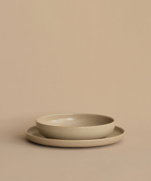 Two round Pacific Dinner Plates are stacked on a matching beige surface and background, with the smaller, deeper plate resting inside the larger, flatter Pacific Dinner Plate.