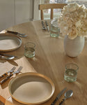 A wooden dining table set for two features Pacific Dinner Plates, glass tumblers, dark-handled cutlery, and a white ceramic vase with pale flowers by a window bathed in natural light.