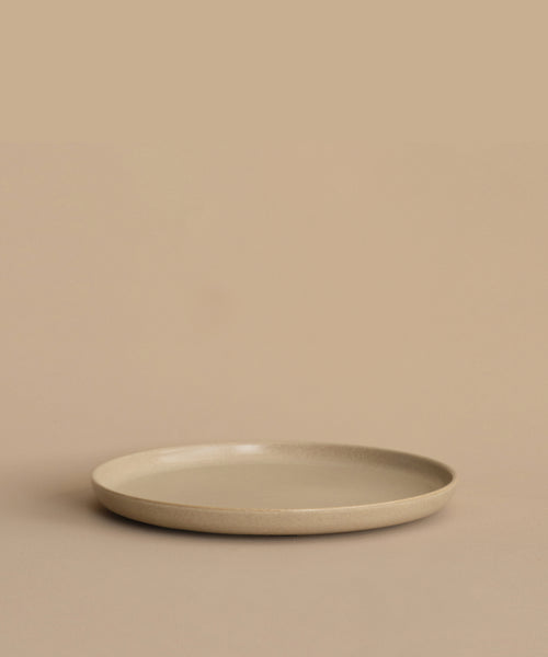The Pacific Dinner Plate, a simple round plate made from beige ceramic, sits on a matching surface with a plain beige backdrop.