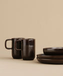 Two Pacific Mugs with handles sit beside two stacked matching plates and a bowl, all arranged on a beige background for a minimalist look.
