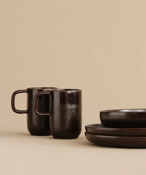 Two Pacific Mugs with handles sit beside two stacked matching plates and a bowl, all arranged on a beige background for a minimalist look.