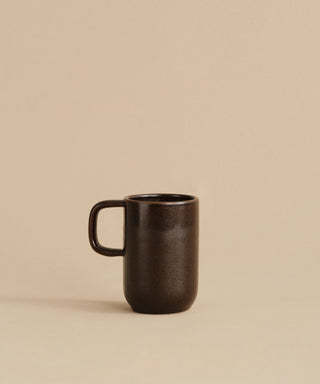 The Pacific Mug, made of dark chocolate-colored stoneware with a handle, rests on a beige surface against a plain beige background.