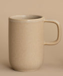 The Pacific Mug, a plain beige stoneware piece with a large handle, rests on a matching surface against a beige background. It features a 10-oz. capacity, blending style and functionality seamlessly.