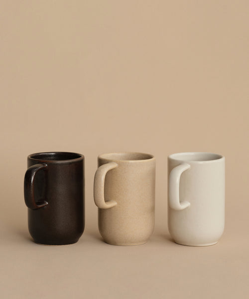 Three Pacific Mugs in dark brown, beige, and off-white are lined up on a beige surface with a matching background. Each Pacific Mug holds 10 oz., ideal for enjoying your favorite beverages.