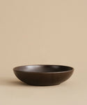 The Pacific Serving Bowl, a stoneware dinnerware piece, sits on a beige surface with a matching background. It features a smooth matte finish and minimalist design in dark brown.