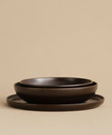 The Pacific Serving Bowl set features three dark brown ceramic pieces of varying sizes—a shallow bowl, a deeper bowl, and a flat plate—stacked together against a beige background. Ideal for adding style to your dinnerware collection.