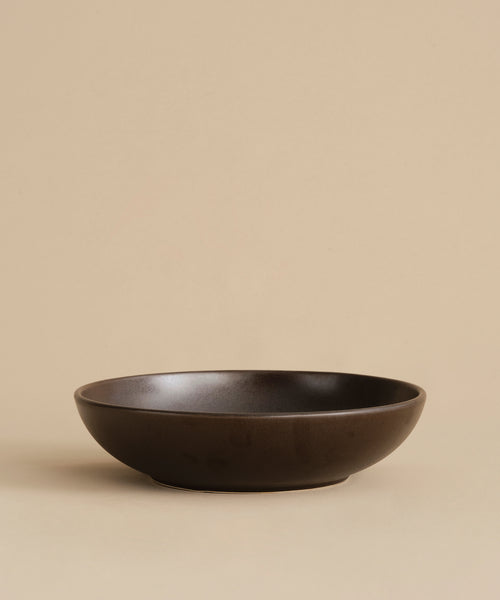 The Pacific Serving Bowl, a stoneware dinnerware piece, sits on a beige surface with a matching background. It features a smooth matte finish and minimalist design in dark brown.