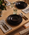 A wooden dining table set with dark brown plates, the Pacific Soup Bowl, glasses of water, beige cloth napkins, cutlery with brown patterned handles, and a flower vase in the corner.