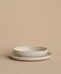 A Pacific Soup Bowl with gently raised sides is stacked on a plain flat plate atop a beige surface. Both white ceramic pieces, made in Portugal, feature a refined and elegant design.