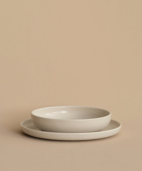 A Pacific Soup Bowl with gently raised sides is stacked on a plain flat plate atop a beige surface. Both white ceramic pieces, made in Portugal, feature a refined and elegant design.