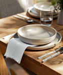 A close-up of a wooden dining table featuring a Pacific Soup Bowl, stoneware dinner plate, white cloth napkin, cutlery with white handles, a glass of water, and a small floral arrangement in sunlight.