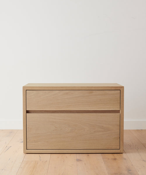 The Palms Nightstand, handmade from solid White Oak, features a simple, modern design with clean lines and no visible handles. Standing on a wooden floor against a white wall, it’s perfect as a nightstand or an accent piece.