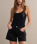 A woman wearing a black sleeveless top and Parker Long Short denim shorts stands with her hands in her pockets against a neutral background.