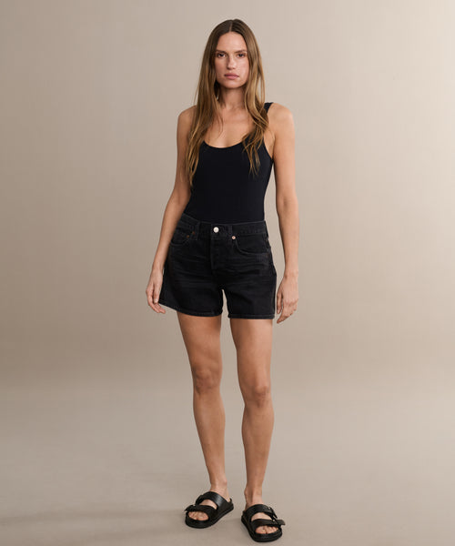 A woman stands against a beige background, wearing a black tank top, Parker Long Short by AGOLDE, and black sandals. She has long straight hair and a neutral expression, with her arms relaxed at her sides.