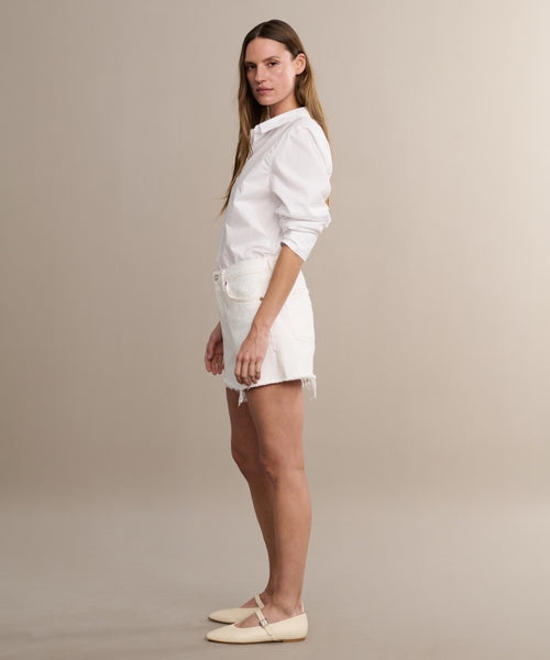 A woman stands in profile against a beige background, wearing a white button-up shirt, Parker Long Short by AGOLDE with frayed hems, and light flat shoes. She has long straight hair and a neutral expression.