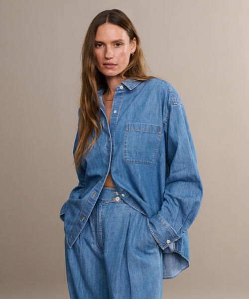 A woman with long brown hair stands with hands in pockets against a beige background, wearing the Parker Oversized Shirt and matching wide-leg denim pants.