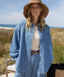 A woman wearing the Parker Oversized Shirt stands outdoors with grass, sand dunes, and blue sky in the background. She pairs the summer-weight denim shirt with a wide-brimmed straw hat, her loose wavy hair framing her neutral expression.